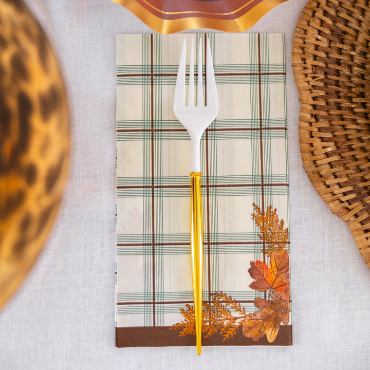 A white fork with a gold handle rests on a plaid napkin from the Sophistiplate Woodland Harvest Guest Towels, adorned with autumn leaves, set atop a white tablecloth beside woven placemats.