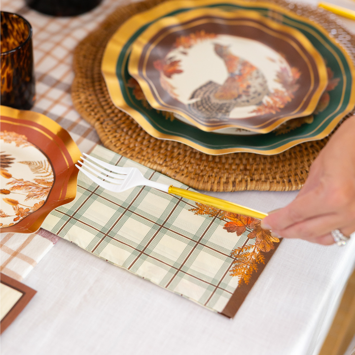 A hand places a gold fork on a plaid napkin next to Sophistiplate’s Woodland Harvest Table Setting, featuring Thanksgiving paper plates with turkey and leaf designs, all arranged in a festive harvest tablescape atop a woven placemat and white tablecloth.
