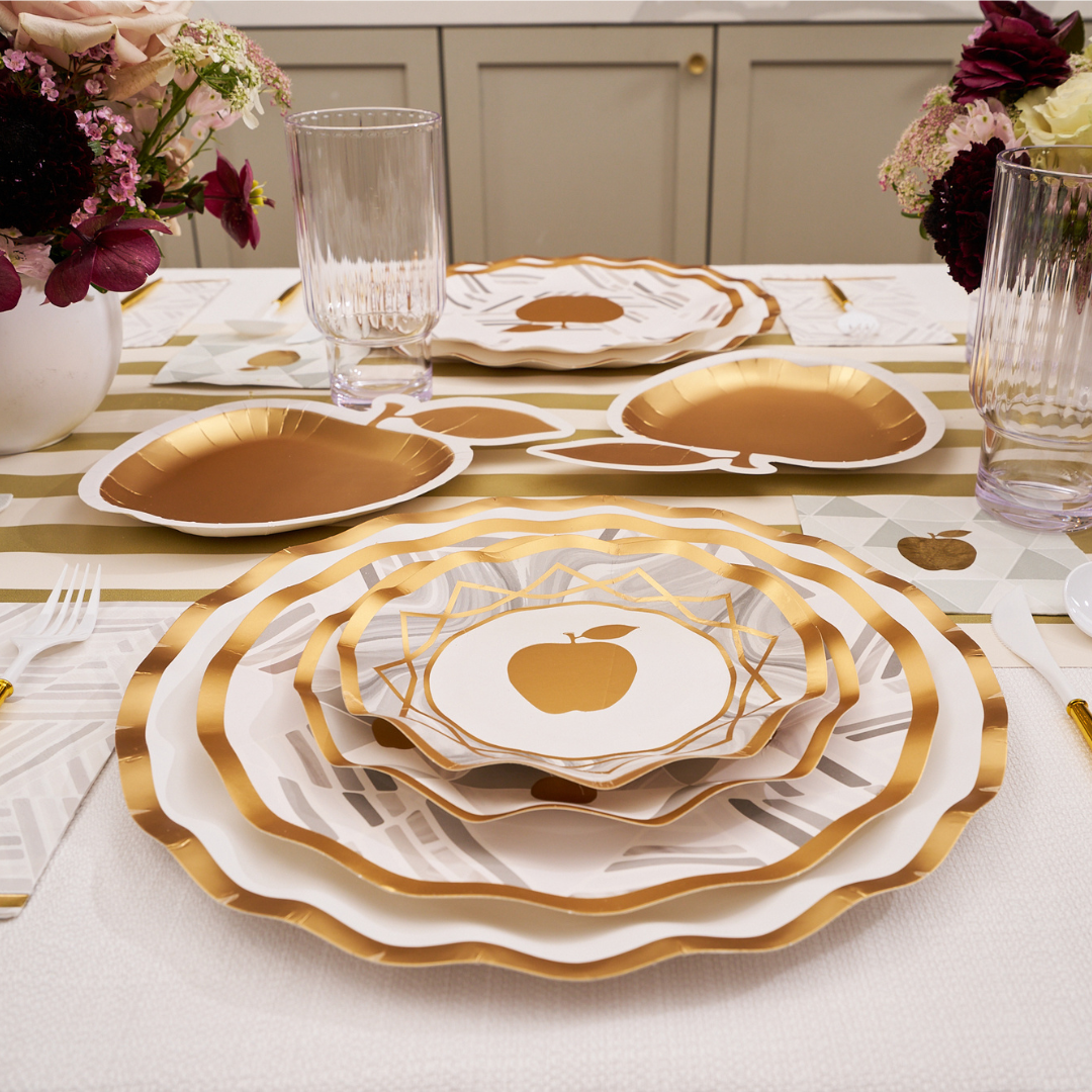 Achieve a festive and stylish table setting for Rosh Hashanah with Sophistiplate's Golden Apple Die Cut Apple Salad Plates (8pk), featuring elegant gold and white designs. Complete the look with golden apple accents, clear glasses, floral centerpieces, and a striped table runner for an elevated holiday presentation.