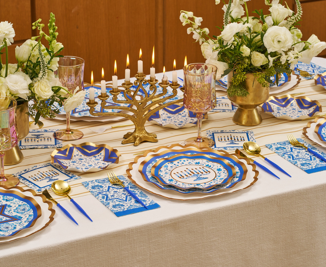 The Hanukkah Toile Table Setting by Sophistiplate features a gold menorah, a festive tablescape with blue and white designer paper plates, gold utensils, blue napkins, and white floral centerpieces atop a cream tablecloth.