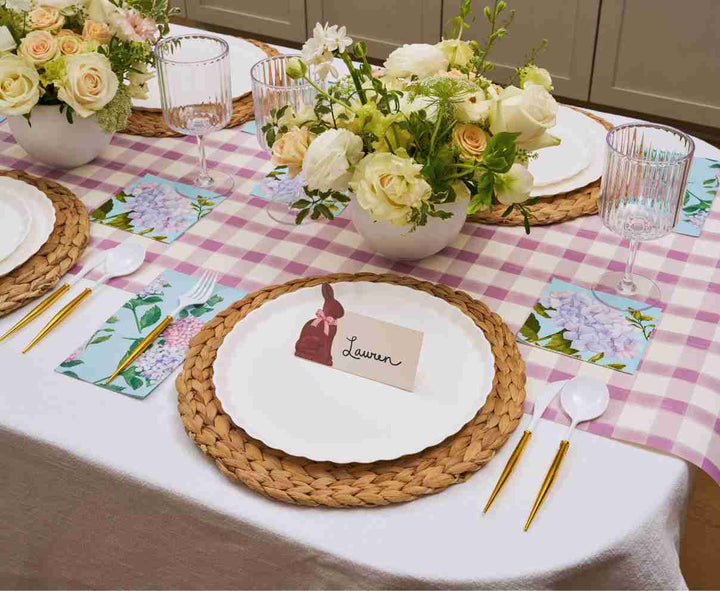 A table set with Sophistiplate’s Ultimate Easter Tablescape for 16 in Violet Hydrangea features designer disposable plates, gold utensils, woven placemats, floral centerpieces, folded napkins with purple flowers, and Lauren's place card with a bunny illustration.