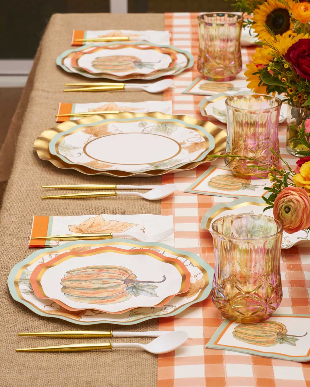 Create a festive autumn tablescape with the Bright & Thankful Table Setting by Sophistiplate, featuring pumpkin-themed plates, gold cutlery, amber glasses, floral napkins, and a sunflower centerpiece on a burlap tablecloth.