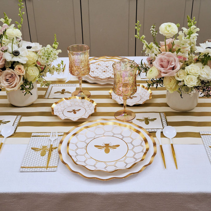 Elevate your celebration with the White Honeybee Table Setting by Sophistiplate, featuring bee-themed gold rim paper plates, gold utensils, a striped gold-and-white runner, pink and gold glasses, and floral centerpieces with roses and ranunculus in white vases—perfect for showcasing elegant party tableware.