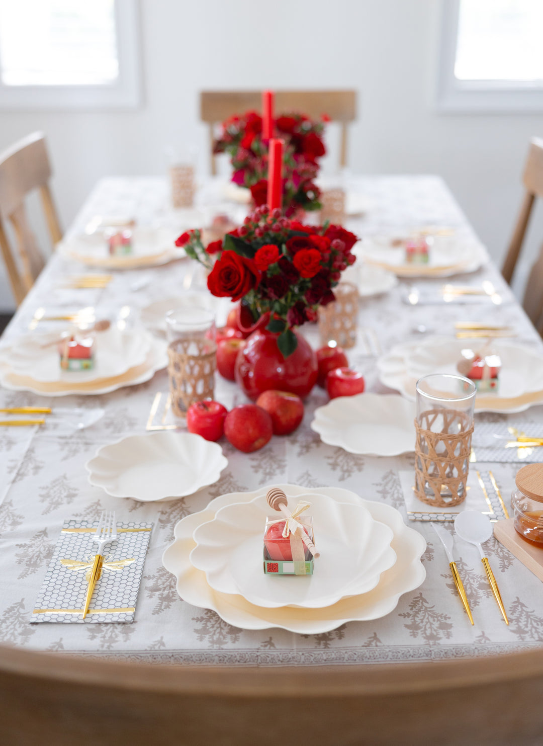 The table is beautifully arranged for Rosh Hashanah with the “Rosh Tablescape for 16 Guests: Eco Cream Honeybee” by Sophistiplate LLC, featuring elegant white plates, gold utensils, glasses, red flowers and pomegranates for a festive touch. Each place setting includes a charming house-shaped favor atop the plate, while two red candles and lush floral centerpieces run down the middle of the table.