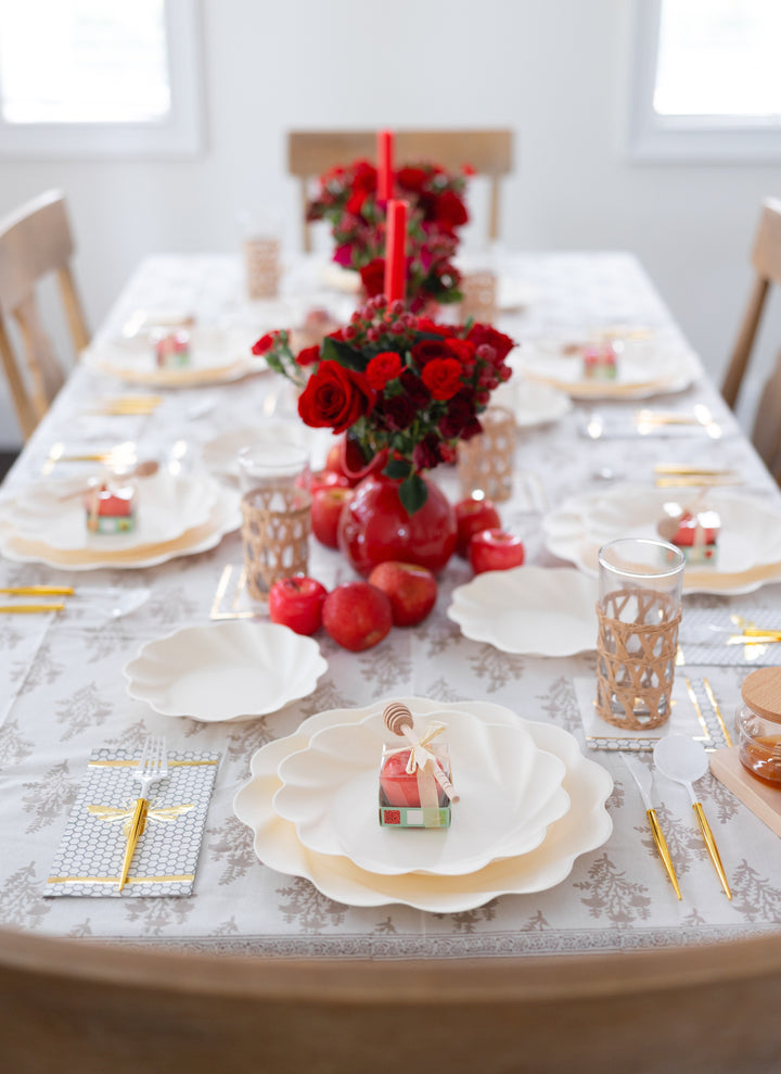 The table is beautifully arranged for Rosh Hashanah with the “Rosh Tablescape for 16 Guests: Eco Cream Honeybee” by Sophistiplate LLC, featuring elegant white plates, gold utensils, glasses, red flowers and pomegranates for a festive touch. Each place setting includes a charming house-shaped favor atop the plate, while two red candles and lush floral centerpieces run down the middle of the table.