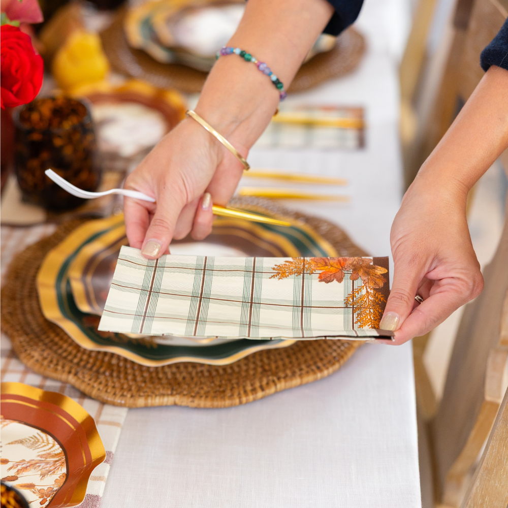 Close-up of hands setting a fall-themed table with a plaid napkin featuring autumn leaves over layered plates, one with a woven charger underneath, alongside Sophistiplate Woodland Harvest Guest Towels/20pk for a festive touch.