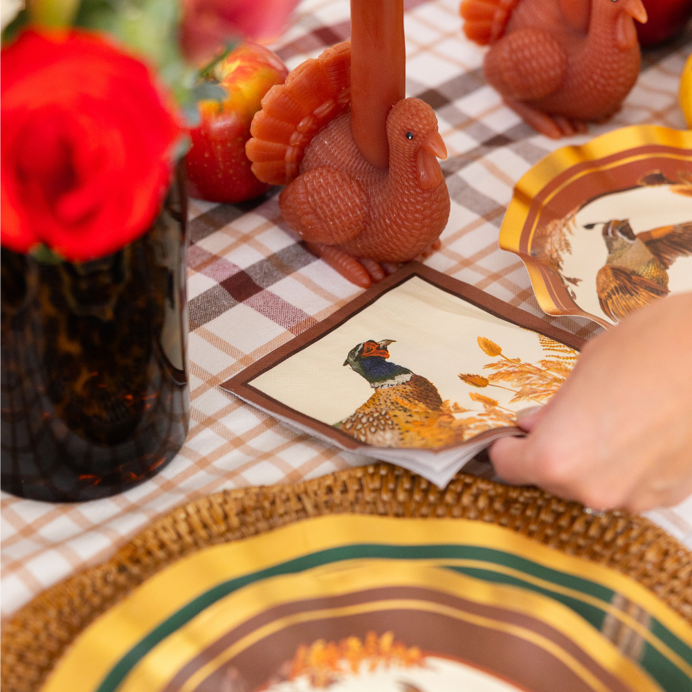 A hand places a Sophistiplate Woodland Harvest Cocktail Napkin from a 26-pack on a plaid tablecloth decorated with turkey-shaped candles, a vase of red flowers, fruit, and a decorative plate, creating a festive autumn table setting.