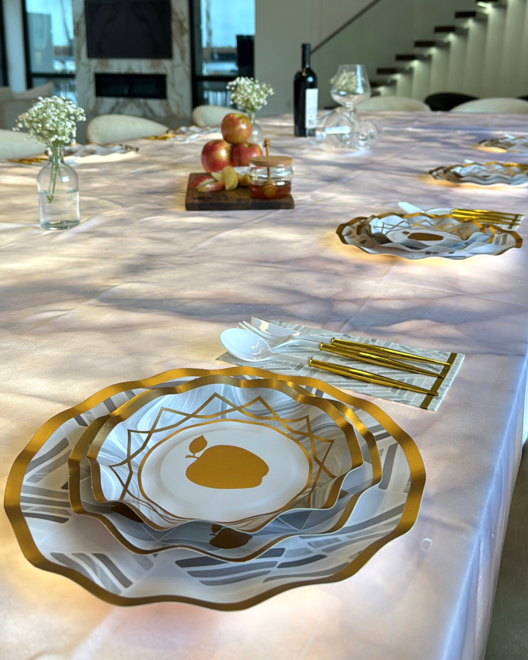 Create a sophisticated Rosh Hashanah table setting with the Sophistiplate LLC Rosh Tablescape for 16 Guests: Golden Apple. This elegant set includes gold-trimmed designer disposable plates, gold utensils, and petite floral vases. A rustic wooden board adorned with apples and a wine bottle rests in the background, while sunlight bathes the scene—perfect for a refined holiday celebration.