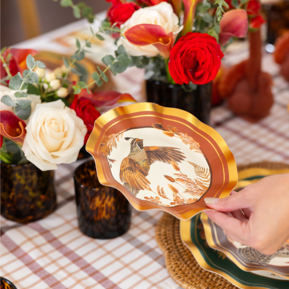 A hand holds a Sophistiplate Woodland Harvest Table Setting designer paper plate featuring a bird motif over a plaid tablecloth. The table is elegantly arranged with floral displays of red and white roses and dark glass tumblers, creating an inviting harvest tablescape.