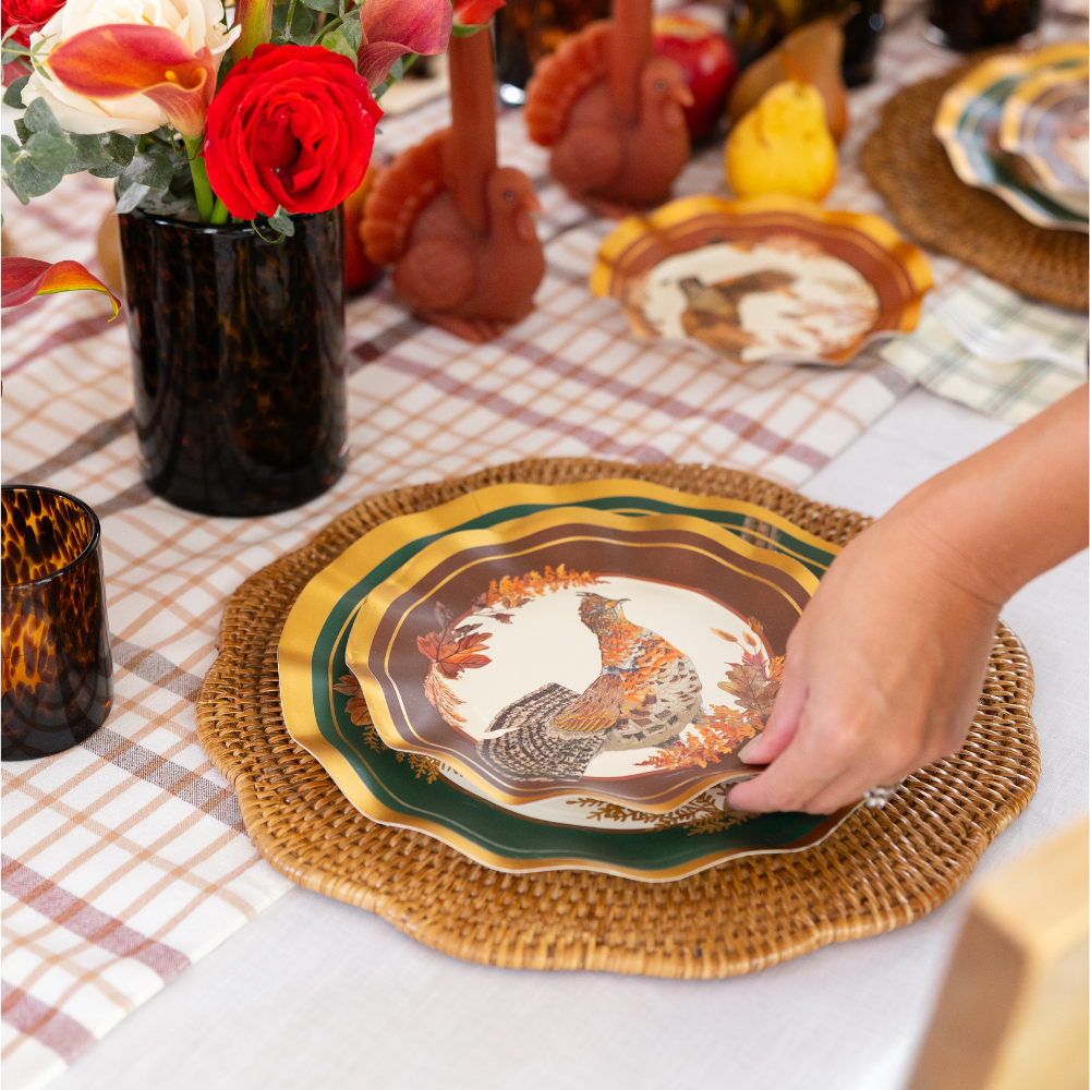 A hand sets a Sophistiplate Woodland Harvest Table Setting paper plate featuring a bird design onto a woven placemat at a festive table, complete with a plaid tablecloth, fresh flowers, and delightful turkey-shaped accents—perfect for elevating your harvest party decor.