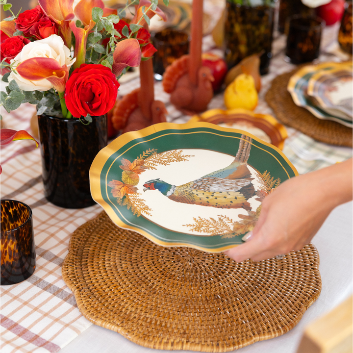 A hand places a Sophistiplate Woodland Harvest Dinner Plate, part of an 8-pack, featuring a pheasant design on a woven placemat atop a checkered tablecloth, surrounded by flowers, candles, and glasses on a festive table.