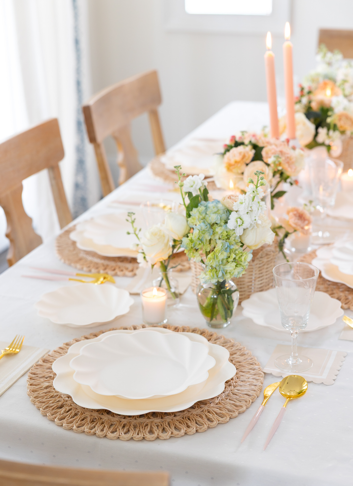 An elegant dining table is set with Sophistiplate Simply Eco Extra Large Plates in Cream, woven placemats, gold and pink cutlery, clear glasses, floral centerpieces, candles, and a white tablecloth for a warm and inviting atmosphere.