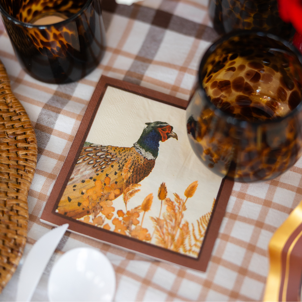 A close-up of a table setting with tortoiseshell glasses and Sophistiplate Woodland Harvest Cocktail Napkins, featuring an illustrated pheasant among autumn plants, on a plaid tablecloth.