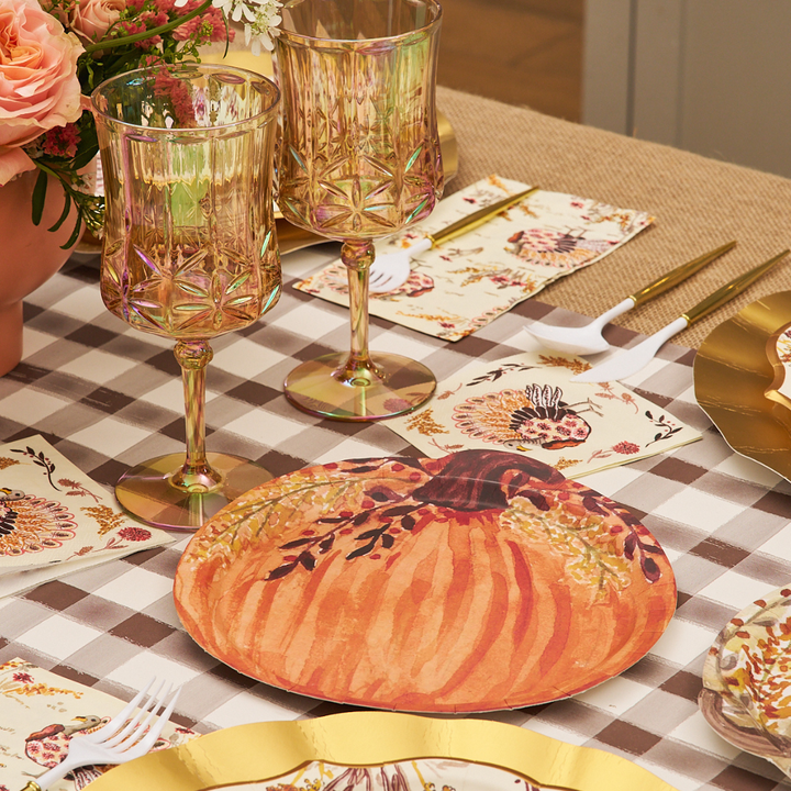 A fall-themed table setting featuring Sophistiplate Amber Harvest Pumpkin Salad Plates, gold utensils, floral napkins, two amber glasses, and pink flowers on a brown and white checkered tablecloth.