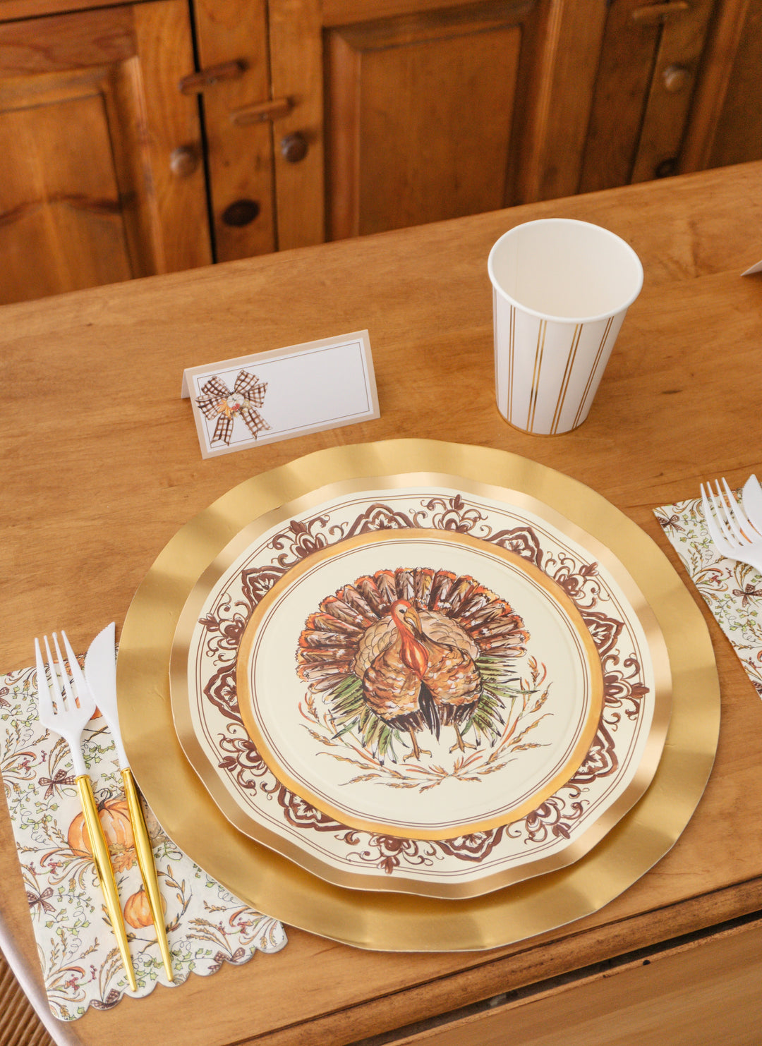 A Sophistiplate LLC Turkey & Gourds: Thanksgiving Table for 16 place setting featuring a decorative turkey heavy duty paper plate on a gold charger, a striped paper cup, a place card with a bow, and a napkin decorated with gourds.
