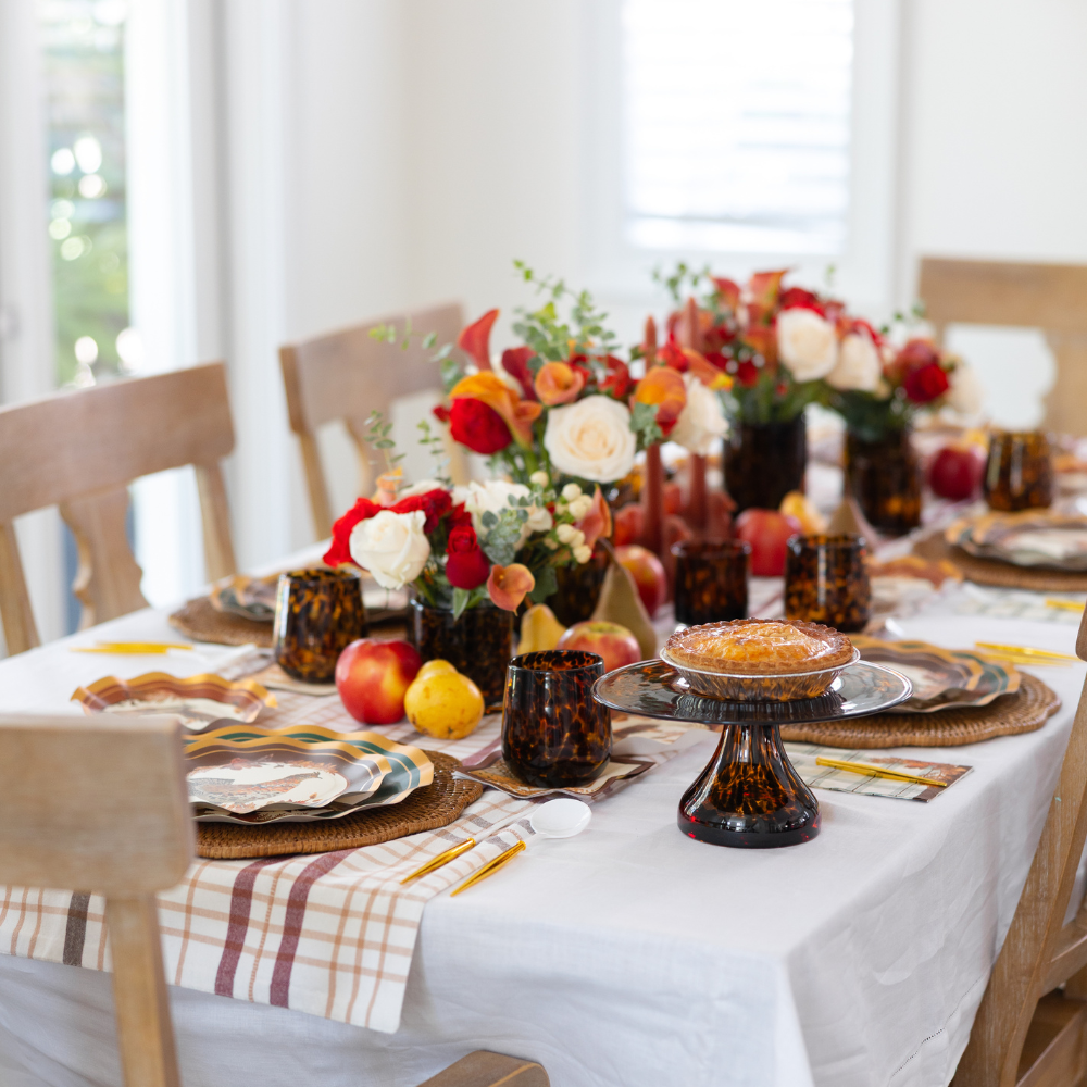 The Woodland Harvest Table Setting by Sophistiplate features a beautifully arranged dining table, complete with floral centerpieces, plaid napkins, amber glassware, harvest-inspired accents, plates topped with pie and fresh apples, all set atop a crisp white tablecloth. Four wooden chairs surround the inviting display.