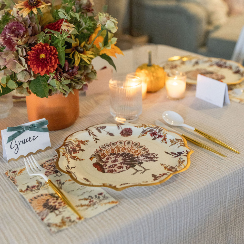 The Amber Harvest Table Setting by Sophistiplate showcases an autumn tablescape with a floral arrangement, candles, a pumpkin, and a decorative plate adorned with a turkey motif. Gold cutlery and a name card labeled Gracee rest on a beige tablecloth alongside the elegant designer disposable plates, creating an inviting seasonal ambiance.