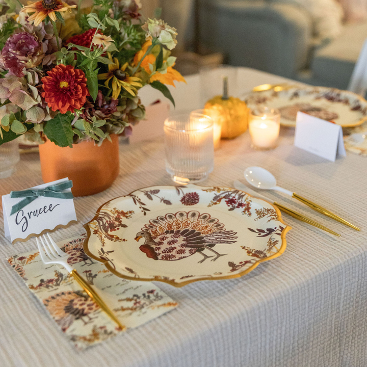 The Amber Harvest Table Setting by Sophistiplate showcases an autumn tablescape with a floral arrangement, candles, a pumpkin, and a decorative plate adorned with a turkey motif. Gold cutlery and a name card labeled Gracee rest on a beige tablecloth alongside the elegant designer disposable plates, creating an inviting seasonal ambiance.
