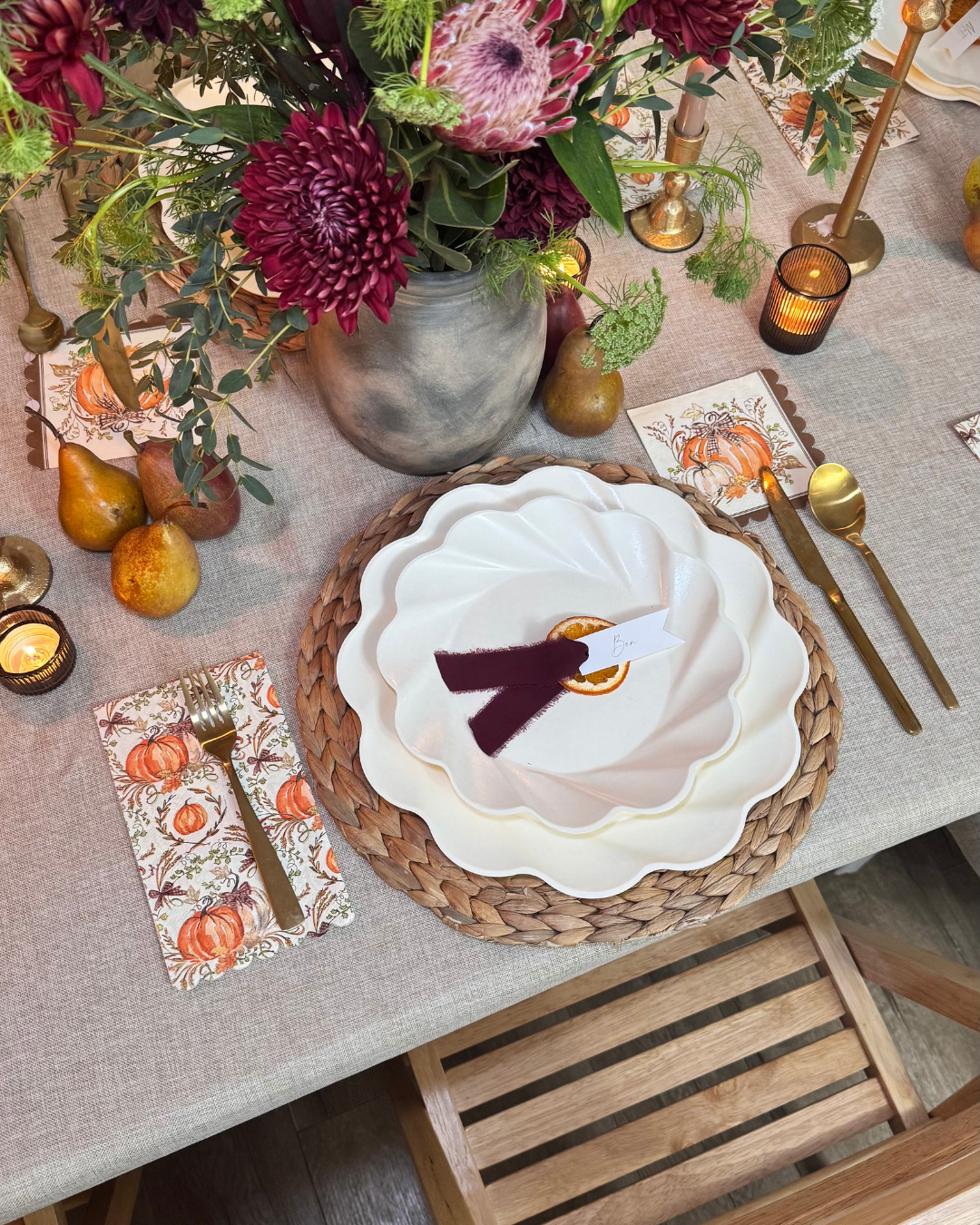 Create a stunning autumn tablescape with the Ivory Harvest Table Setting by Sophistiplate, showcasing eco-friendly disposable plates or classic white plates set atop woven chargers. Complement the look with gold cutlery, autumn-themed napkins, ripe pears, glowing candles, and a vibrant floral centerpiece featuring red and pink blooms on a beige tablecloth—perfect for highlighting Thanksgiving dinnerware in sustainable style.