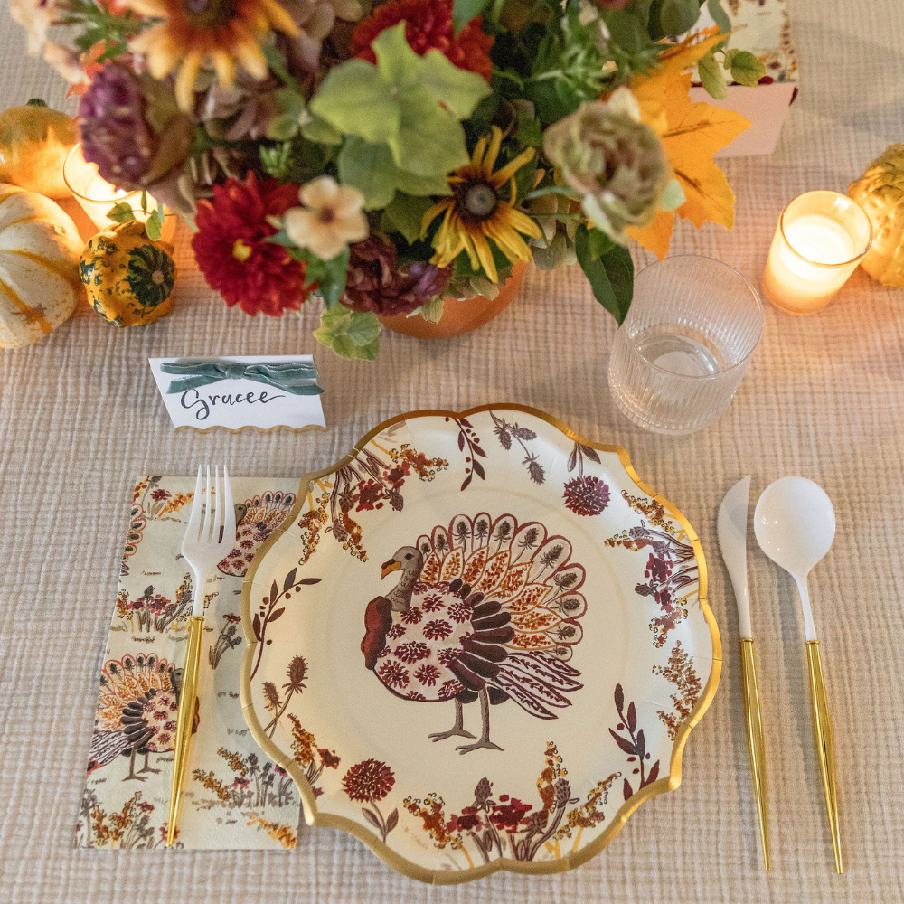 The Amber Harvest Table Setting by Sophistiplate features a decorative turkey plate from our designer disposable plates collection, gold utensils, a floral napkin, a glass, a lit candle, a bouquet of flowers, small pumpkins, and a place card labeled Gracee—creating the perfect harvest tablescape for your gathering.