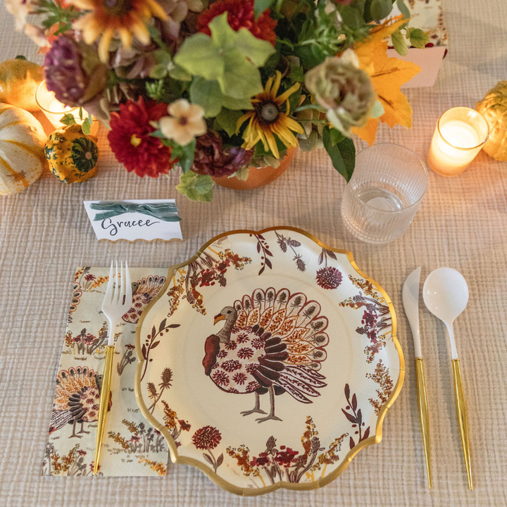 The Amber Harvest Table Setting by Sophistiplate features a decorative turkey plate from our designer disposable plates collection, gold utensils, a floral napkin, a glass, a lit candle, a bouquet of flowers, small pumpkins, and a place card labeled Gracee—creating the perfect harvest tablescape for your gathering.