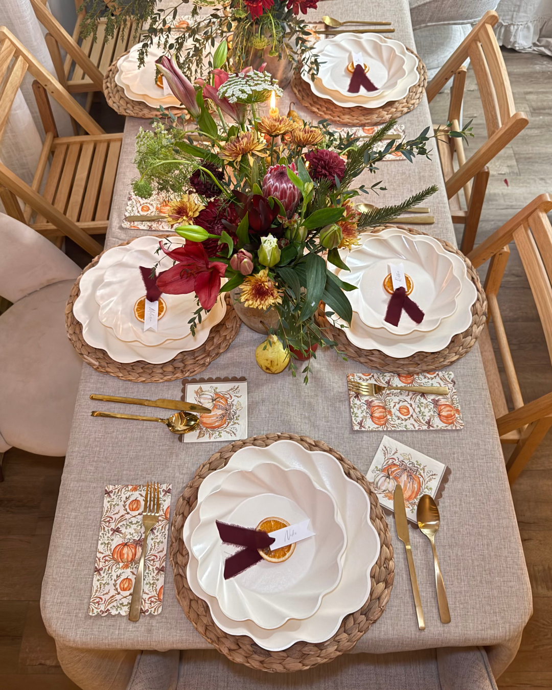 The dining table is elegantly set for six with the Sophistiplate Ivory Harvest Table Setting, featuring Thanksgiving-themed tableware, gold cutlery, autumn-inspired napkins, and white scalloped plates. At the center is a vibrant display of red, yellow, and green flowers. Each plate includes a ribbon-tied tag, adding a festive and refined touch to the décor.