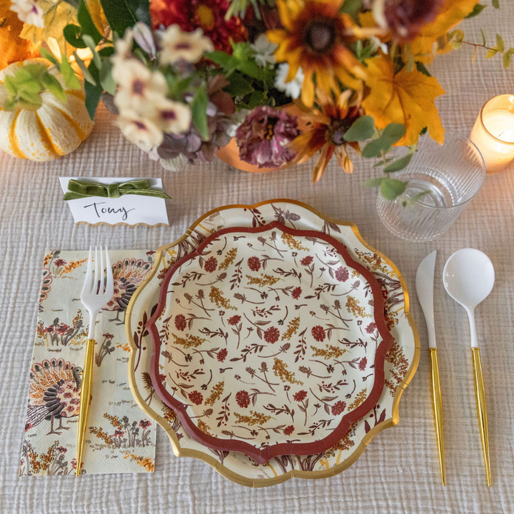 Create a warm autumn mood with the Amber Harvest Table Setting by Sophistiplate, featuring designer disposable plates adorned with florals, elegant gold cutlery, a turkey-themed napkin, a personalized name card for Tony, sparkling glassware, a glowing candle, and a vibrant bouquet of fall flowers—all set atop a rich textured tablecloth.