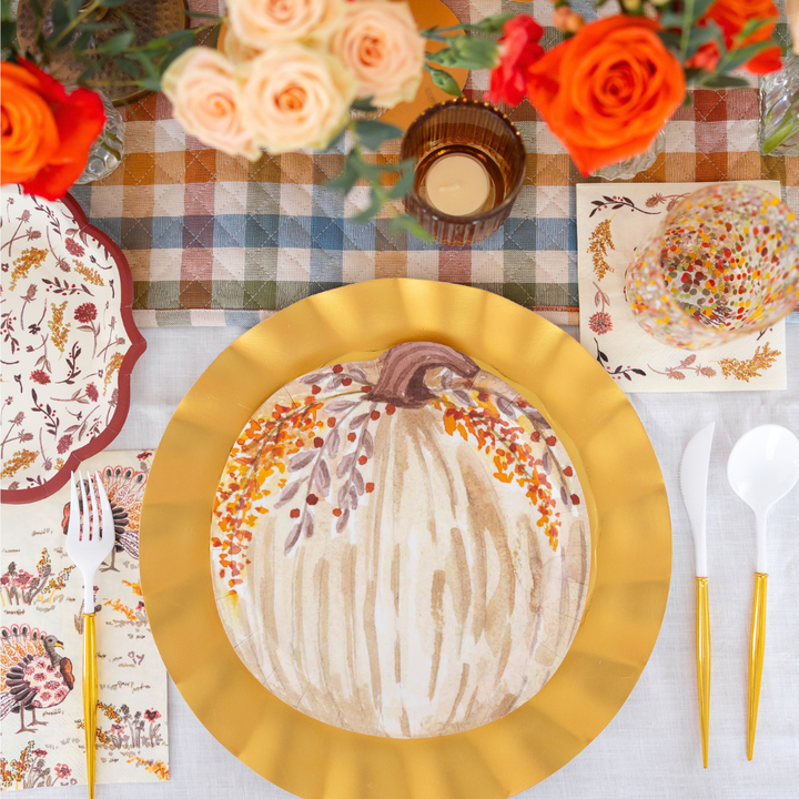 Create a festive table with the Sophistiplate Amber Harvest Table Setting, featuring gold-edged plates adorned with a pumpkin design, autumn-themed napkins, plastic cutlery, a candle, and vibrant florals on a plaid tablecloth—an elegant harvest tablescape perfectly finished with designer disposable plates and other autumn party supplies.