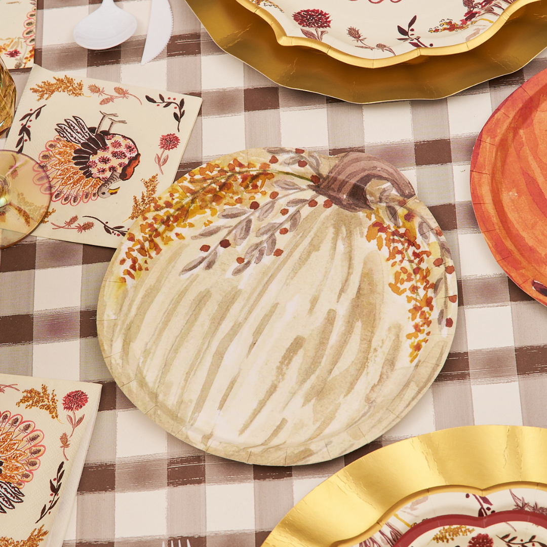 Round paper plate from the Sophistiplate Amber Harvest Pumpkin Salad Plate (8pk), featuring an autumn pumpkin and leaves design on a brown checkered tablecloth, surrounded by gold-trimmed disposable plates and napkins adorned with turkeys and fall foliage patterns.