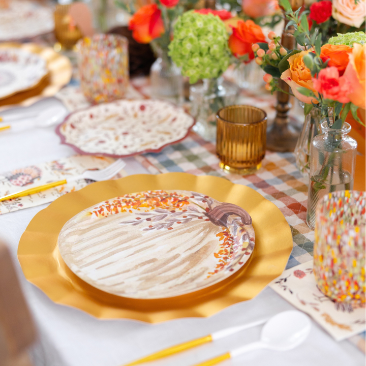 Create a stunning autumn tablescape with the Amber Harvest Table Setting by Sophistiplate, featuring designer disposable plates and gold chargers, amber glasses, floral centerpieces in orange and green hues, and gold cutlery arranged on a plaid tablecloth—perfect for festive gatherings.