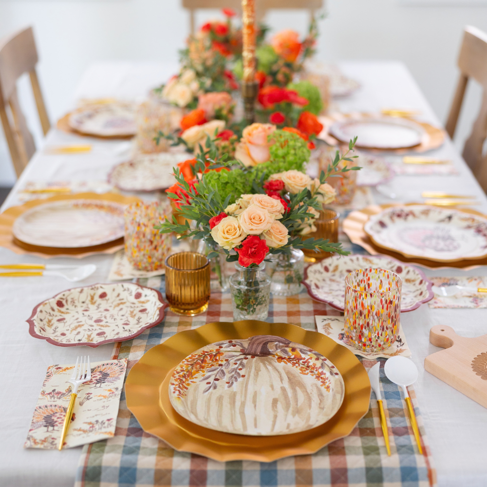 Celebrate the season with the Amber Harvest Table Setting by Sophistiplate—a festive dining arrangement featuring autumn-themed plates adorned with pumpkin and leaf designs, elegant gold flatware, floral centerpieces, and a stylish checkered tablecloth. Vibrant cups and neatly arranged napkins complete this designer disposable set, making it perfect for highlighting your autumn party supplies.