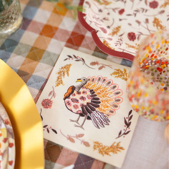 Elevate your autumn celebration with the Amber Harvest Table Setting from Sophistiplate, featuring a gold charger, elegant designer disposable plates with intricate patterns, a glass embellished with colorful dots, and a napkin displaying a festive turkey amid autumn leaves—all beautifully arranged on a plaid tablecloth.