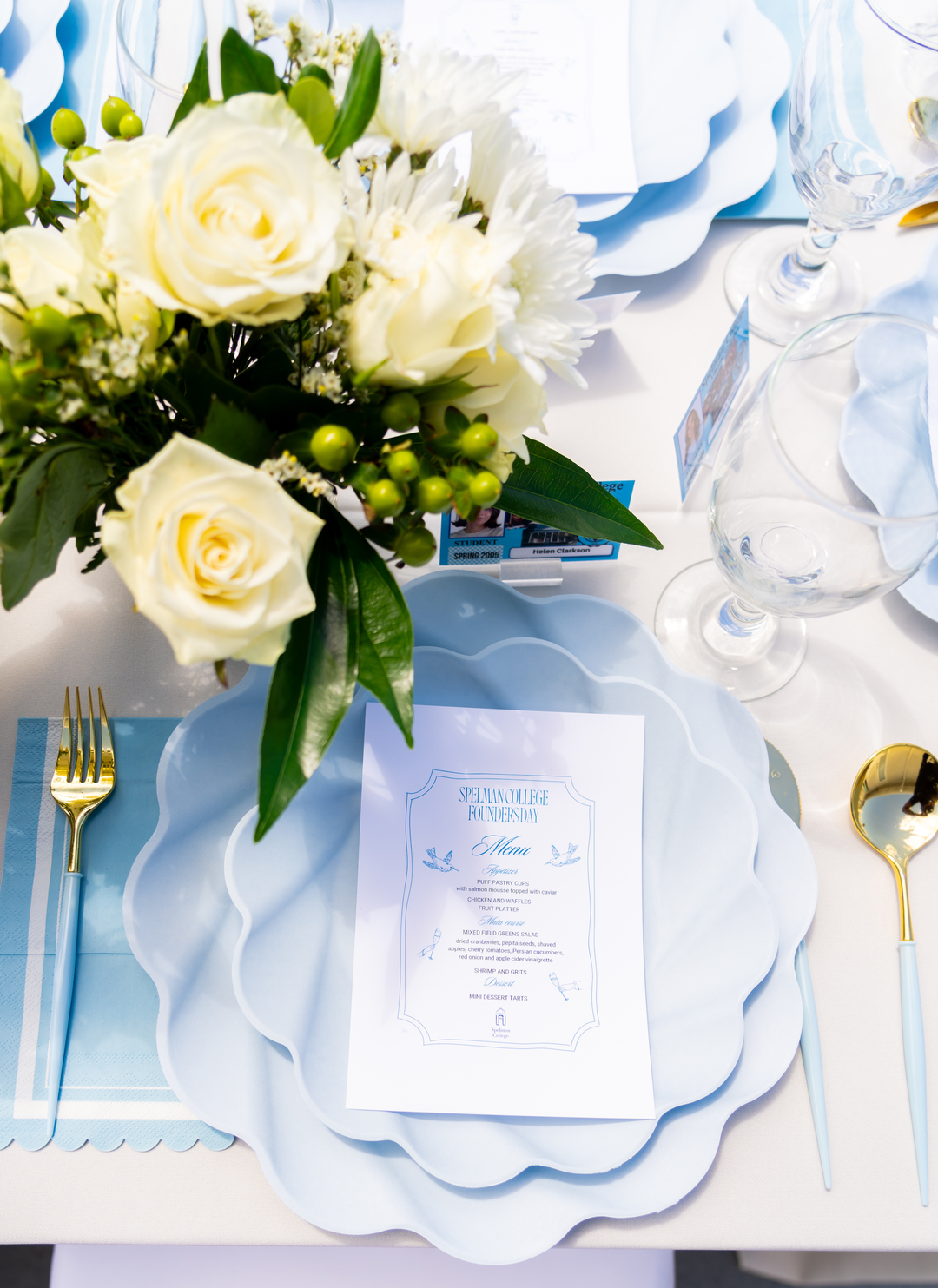 A close-up of an elegant table setting with Sophistiplate's Sky Blue Scalloped Edge Guest Towels, a white menu card, gold flatware, clear glassware, eco-friendly tableware, compostable napkins, and a white rose and greenery centerpiece.