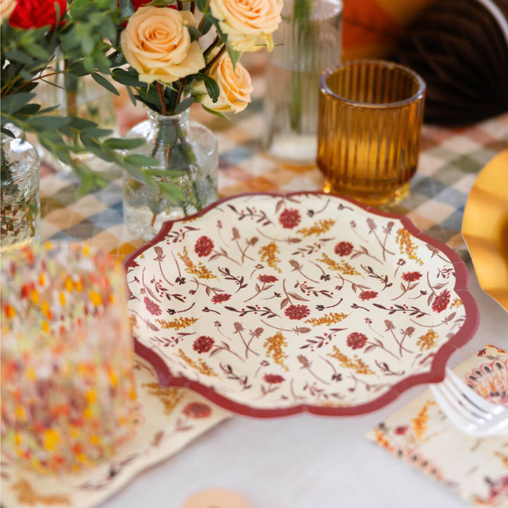 The Amber Harvest Table Setting by Sophistiplate features a floral-patterned designer disposable plate arranged on a set table with a yellow glass, multicolored tumbler, silverware, and vases of yellow and peach roses atop a checkered tablecloth—perfect for creating an inviting autumn tablescape.