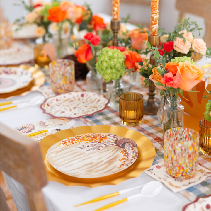 Create a warm and inviting autumn or Thanksgiving atmosphere with the Amber Harvest Table Setting by Sophistiplate, featuring gold chargers, patterned designer disposable plates, orange and yellow floral arrangements, amber glasses, and candles for a stunning harvest tablescape.