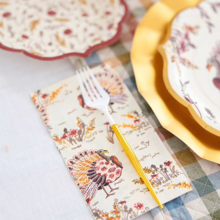 The Amber Harvest Table Setting by Sophistiplate creates a festive autumn tablescape with turkey-themed napkins, a yellow-handled fork, and vibrant harvest party plates, all beautifully arranged on a cheerful plaid tablecloth.