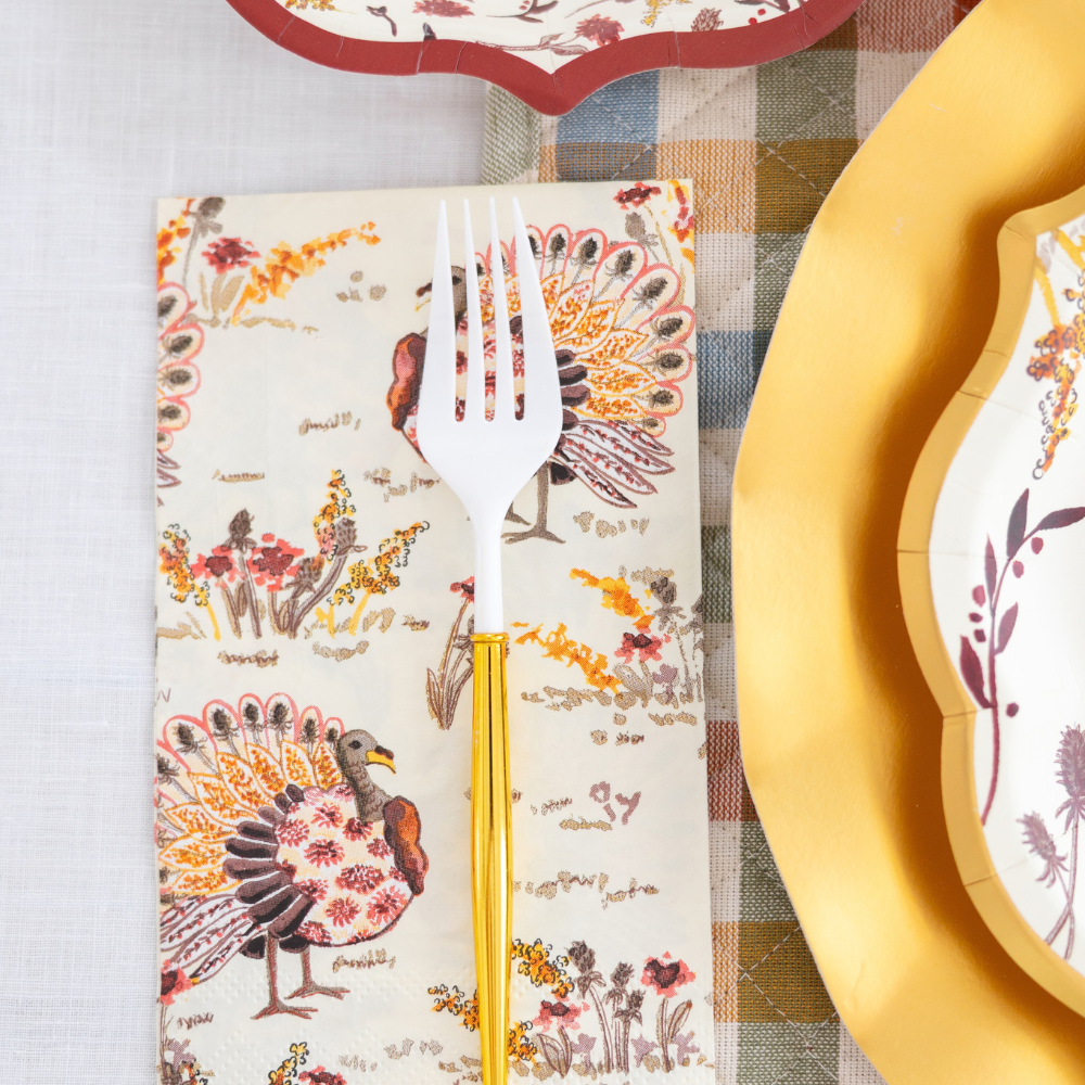 The Amber Harvest Table Setting by Sophistiplate features a white fork with a gold handle resting on a napkin adorned with colorful turkeys, accompanied by designer disposable plates and a patterned plate atop a plaid tablecloth, creating an inviting autumn tablescape.