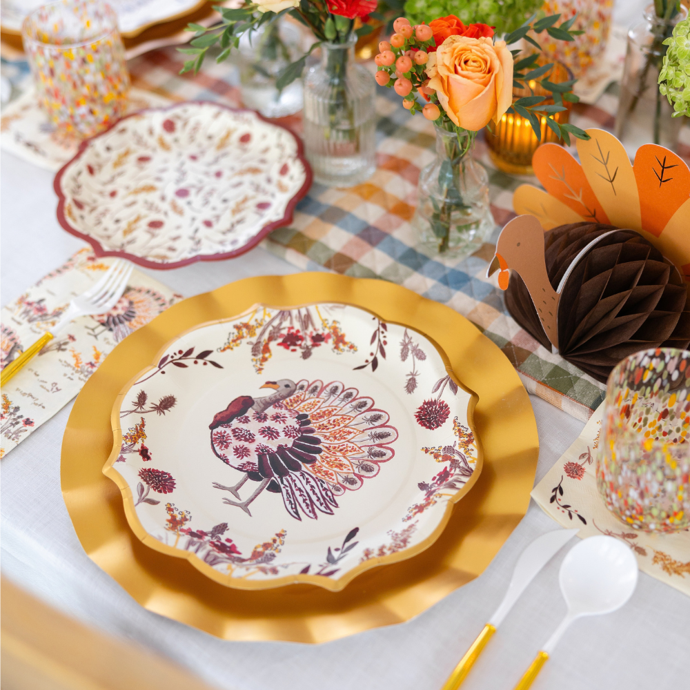 The Amber Harvest Table Setting by Sophistiplate features gold-rimmed chargers, harvest party plates, patterned napkins and glasses, a paper turkey decoration, and orange flowers arranged on a plaid tablecloth to create an inviting autumn tablescape for Thanksgiving.