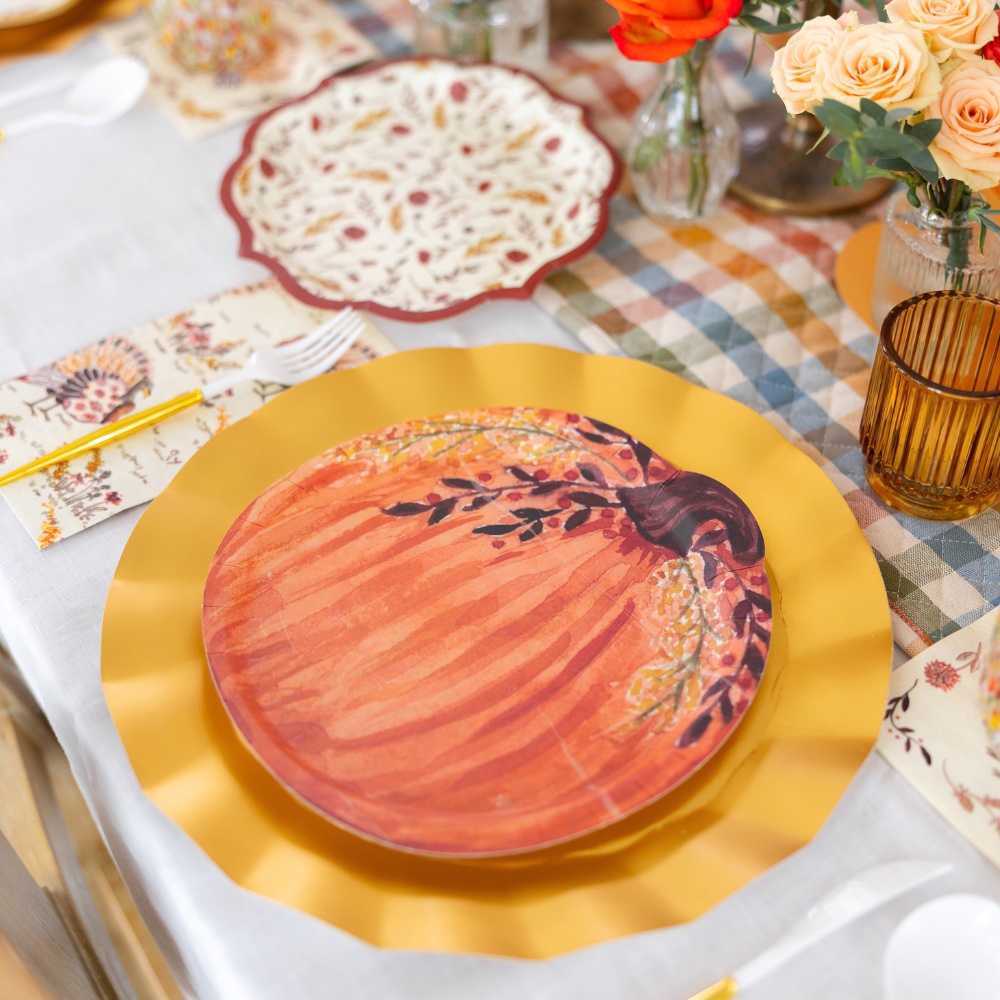Create a festive autumn table with the Amber Harvest Table Setting by Sophistiplate, featuring a gold charger, pumpkin-themed harvest party plates, a floral napkin, cutlery, amber glassware, and small vases filled with orange flowers artfully arranged atop a plaid tablecloth.