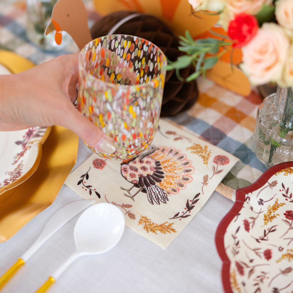 A hand holds a colorful glass over a Sophistiplate Amber Harvest Cocktail Napkin on a festive table set with floral plates, plastic cutlery, flowers, and autumn decorations.