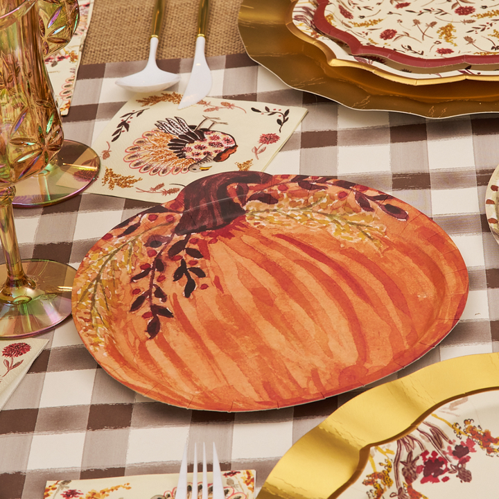 Create a festive table setting with the Sophistiplate Amber Harvest Pumpkin Salad Plates (8-pack), paired with gold-rimmed glasses, a floral napkin featuring a turkey design, and patterned plates atop a brown and white checkered tablecloth. Autumn-themed decor sets the scene, while the charming pumpkin-shaped salad plates from Sophistiplate add a delightful seasonal touch perfect for any harvest party.
