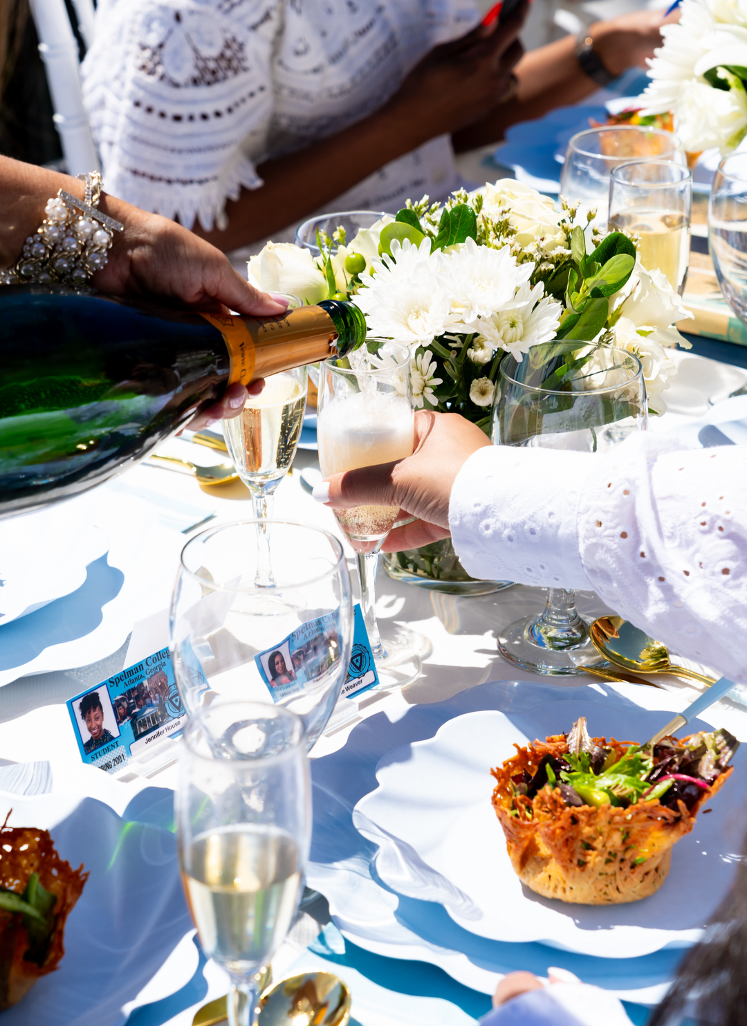 A person pours champagne at an elegantly set table with white flowers, salads, and Sophistiplate Simply Eco Dinner Plate- Sky Blue/8pkg as guests in white enjoy an outdoor meal.