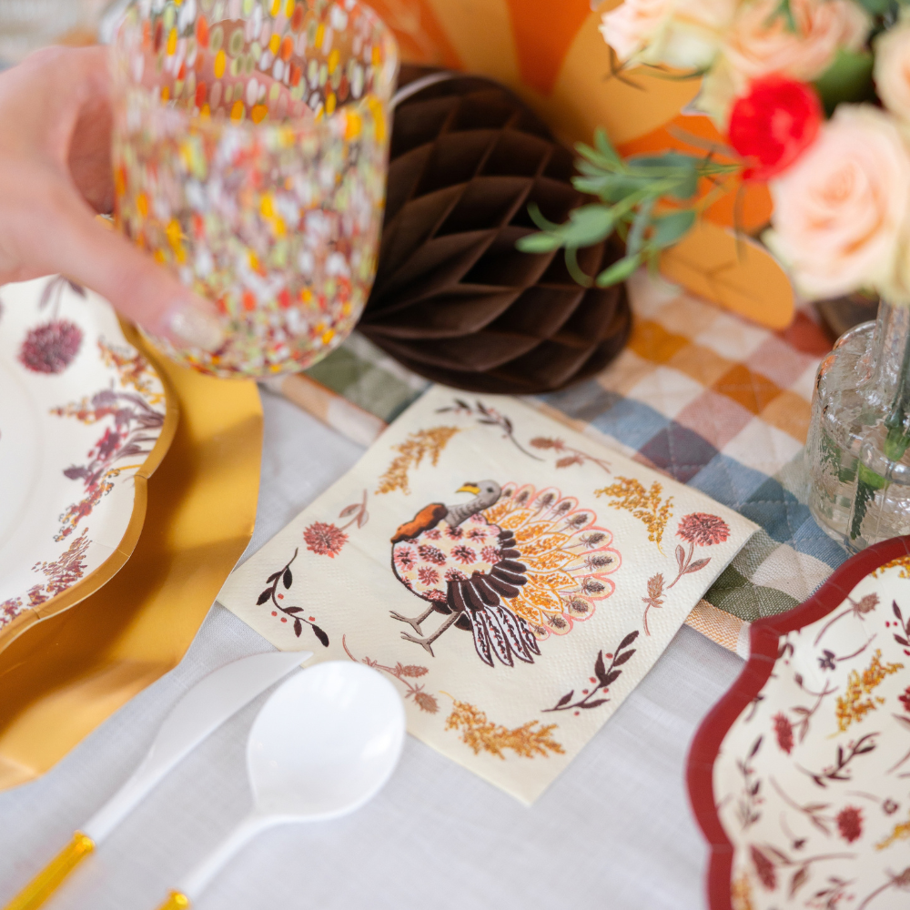 A hand holds a colorful glass above a table accented with Sophistiplate’s Amber Harvest Table Setting, featuring floral designer disposable plates, white cutlery, a turkey-themed napkin, and a vase of roses.