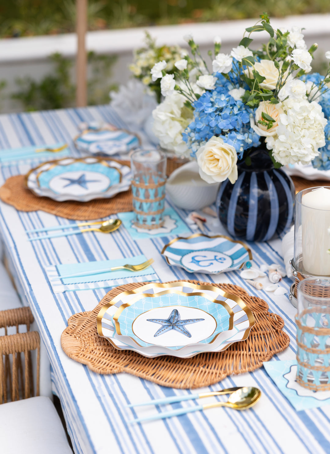 Create a festive coastal tablescape with the Tidal Wave Table Setting by Sophistiplate—featuring starfish plates, woven chargers, blue and white stripes, gold utensils, rattan glasses, a floral centerpiece, and scattered seashells.