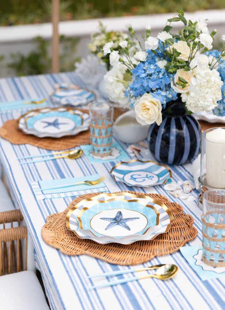 Create a festive coastal tablescape with the Tidal Wave Table Setting by Sophistiplate—featuring starfish plates, woven chargers, blue and white stripes, gold utensils, rattan glasses, a floral centerpiece, and scattered seashells.