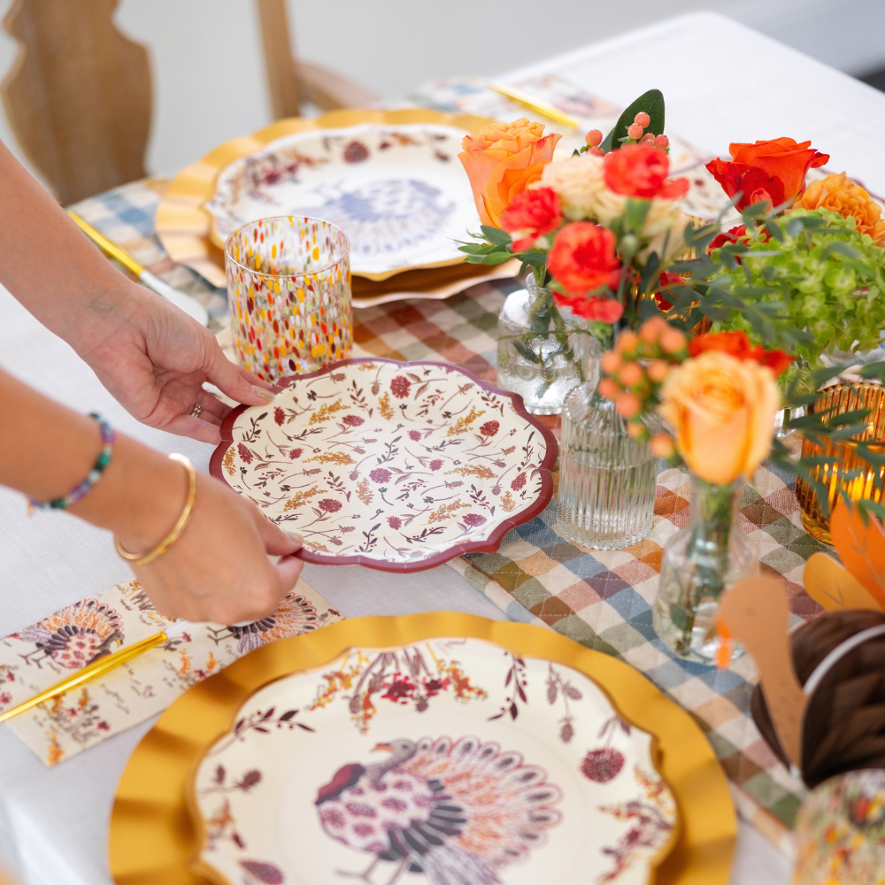 A person sets a Sophistiplate Amber Harvest Table Setting on a festive table adorned with orange and yellow flowers and a colorful checked table runner, creating a warm autumn tablescape inspired by the season.