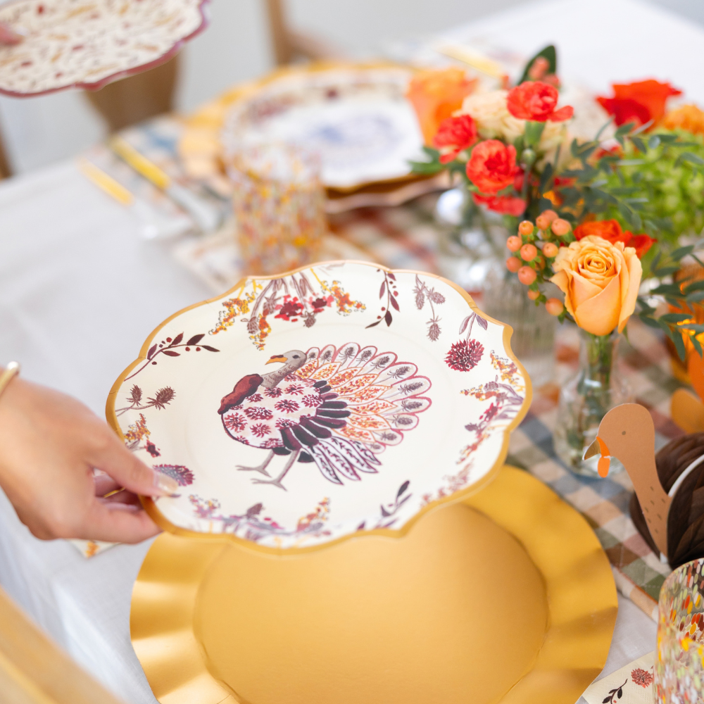 A hand places a decorative plate featuring a turkey design—part of the Amber Harvest Table Setting by Sophistiplate—on a gold charger at a table adorned with orange flowers, plaid napkins, and festive Thanksgiving decor, creating the perfect autumn tablescape.