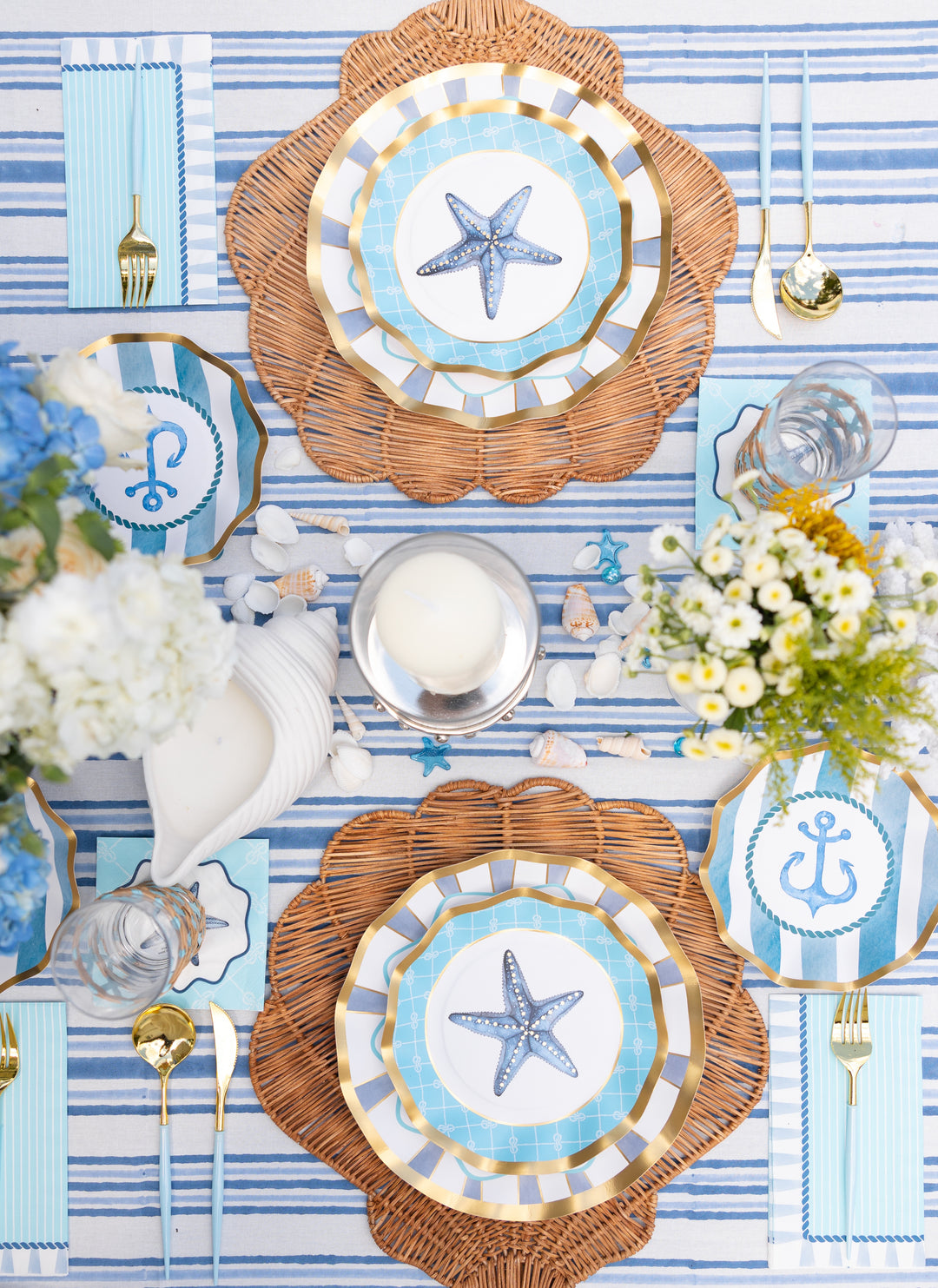 Create a coastal-themed party look with the Sophistiplate Tidal Wave Table Setting, featuring wicker placemats, gold cutlery, striped napkins, and designer paper plates with starfish and anchor motifs on a blue and white striped tablecloth.