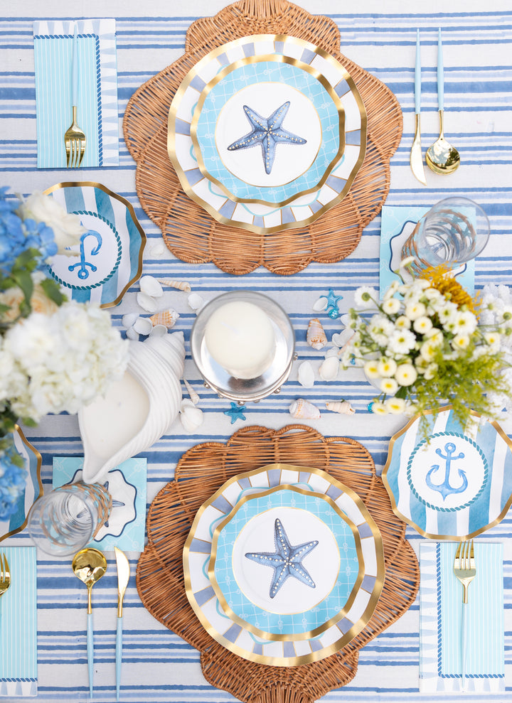 Create a coastal-themed party look with the Sophistiplate Tidal Wave Table Setting, featuring wicker placemats, gold cutlery, striped napkins, and designer paper plates with starfish and anchor motifs on a blue and white striped tablecloth.