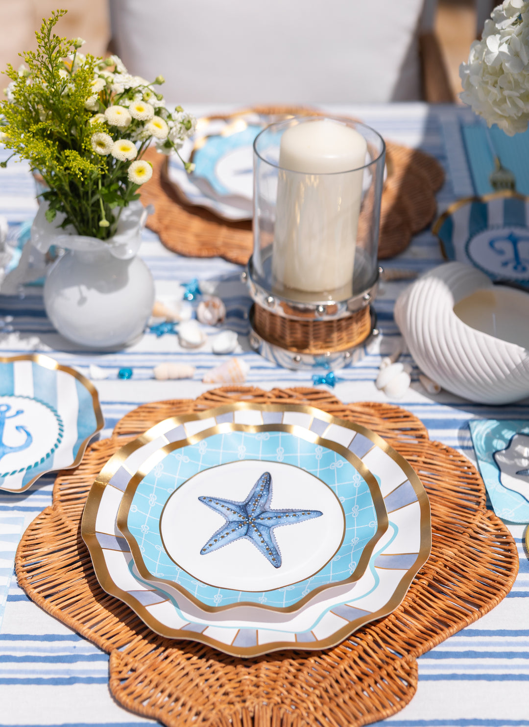 The Tidal Wave Table Setting by Sophistiplate features coastal starfish plates, gold accents, a wicker placemat, glass candle holder, white flowers in a vase, and seashells atop a striped tablecloth for a nautical-themed party.